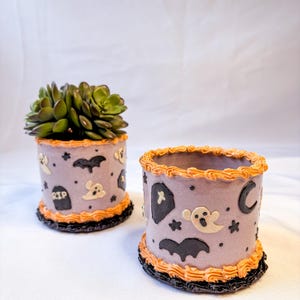 May include: Two Halloween-themed cylindrical planters. One holds a green succulent. Both are lavender with orange and black trim. Decorations include ghosts, bats, stars, and a tombstone with "RIP". The other planter has a crescent moon and coffin.