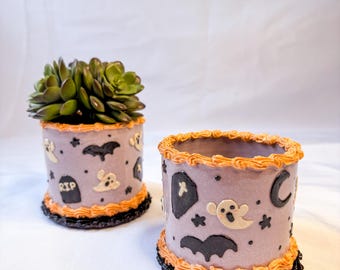 以下が含まれることがあります： ハロウィーンをテーマにした2つの円筒形のプランター。1つは緑色の多肉植物が入っています。どちらもラベンダー色で、オレンジと黒のトリムが付いています。装飾には、幽霊、コウモリ、星、そして「RIP」と書かれた墓石が含まれています。もう一方のプランターには三日月と棺があります。