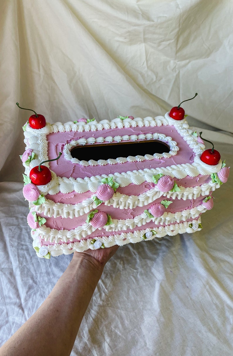 May include: A pink and white tissue box shaped like a cake with white frosting and red cherries on top.