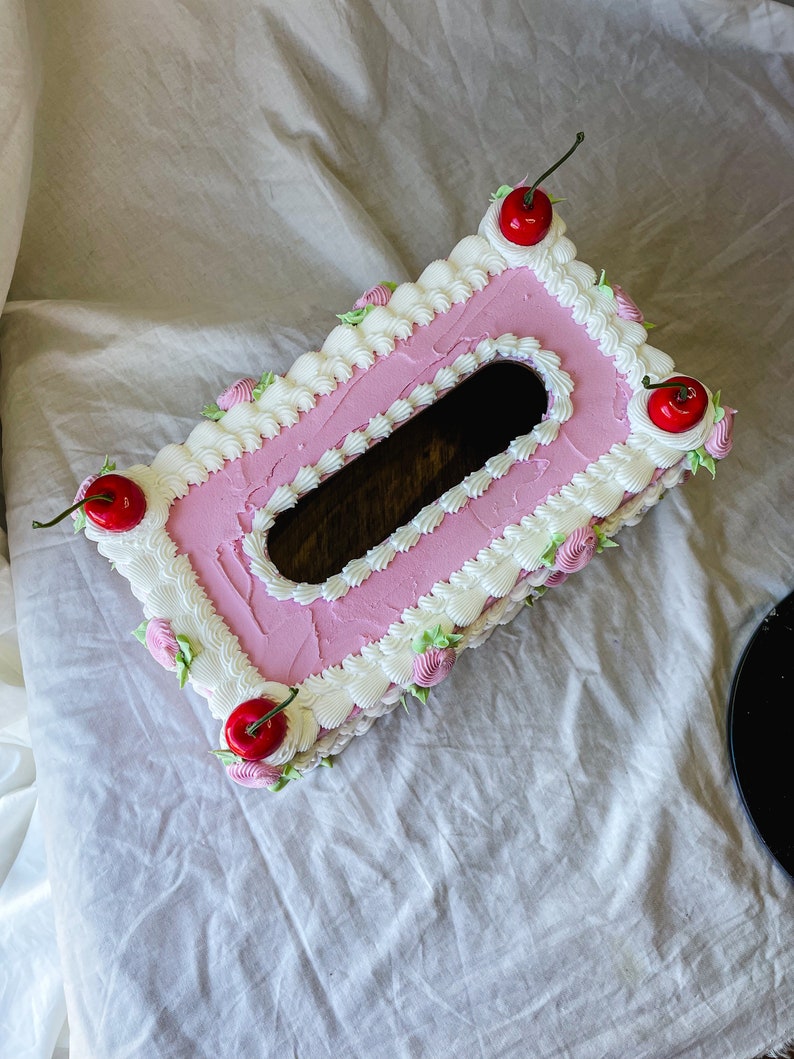 May include: A pink and white tissue box cover shaped like a cake with white frosting and red cherries on top.