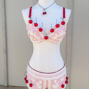 May include: A pink lingerie set with cherry accents, displayed on a mannequin. The set includes a bra, shorts, and a necklace, all adorned with red cherries. The bra and shorts feature white lace detailing.
