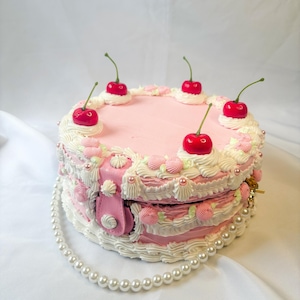 May include: A pink cake-shaped handbag with white frosting, pink roses, and red cherries. The handbag has a pearl strap and a pink clasp. The cake-shaped handbag is a unique accessory.