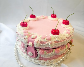 May include: A pink cake-shaped handbag with white icing, pink roses, and red cherries. The handbag has a pearl strap and a pink clasp. The cake-shaped handbag is a unique accessory.