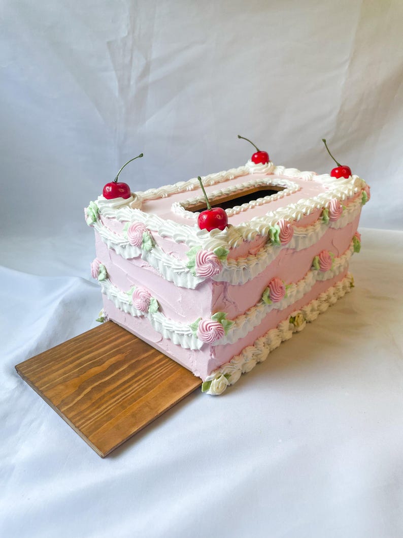 May include: A pink and white cake-shaped tissue box cover with red cherries on top. The box has a wooden base that slides out.