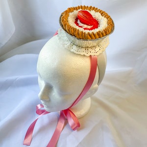May include: A pink ribbon headband with a miniature pie on top. The pie has a brown crust, white frosting, and a red strawberry filling.