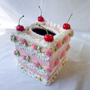 May include: A pink and white tissue box cover shaped like a cake with white frosting and pink roses. The cake is decorated with four red cherries on top.