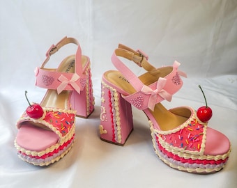May include: Pink platform sandals with a cake-like design. The sandals feature a cherry on the toe, a pink bow, and a heart-shaped embellishment. The heels are square and decorated with white frosting-like details.