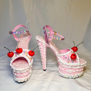 May include: Pink platform high heels with a cake-like design. The heels feature white frosting details, colorful sprinkles, and red cherry accents. The ankle straps are iridescent.