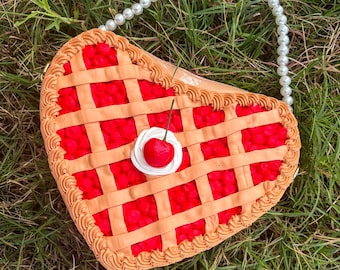 May include: A novelty handbag shaped like a heart-shaped cherry pie. The bag has a red filling, lattice crust, and a cherry on top. It is finished with a pearl beaded handle.