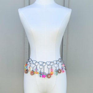 May include: A silver-toned belt with heart-shaped links and dangling charms. The charms include colorful candies, cookies, and lollipops. The belt is displayed on a white mannequin, showcasing its design and details.