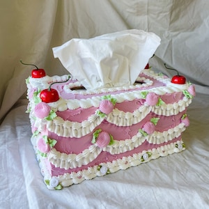 May include: A pink tissue box shaped like a cake with white frosting and green and pink decorations. There are three red cherries on top of the cake.