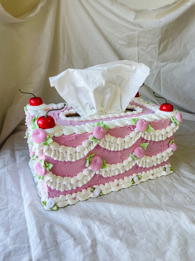 May include: A pink tissue box shaped like a cake with white frosting and green and pink decorations. There are three red cherries on top of the cake.