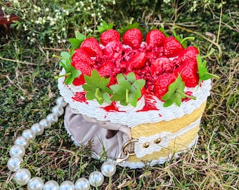 May include: A novelty cake-shaped handbag with a pearl strap. The bag features a yellow and white cake design, topped with red strawberries and green leaves. The strap is made of large, round, white pearls.