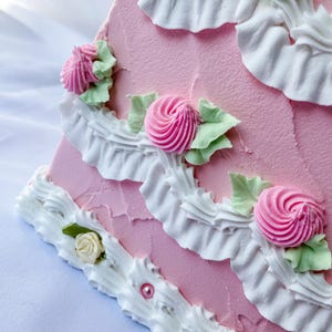 May include: A pink cake decorated with white frosting and pink and white flowers. The cake is layered and has a ruffled edge.
