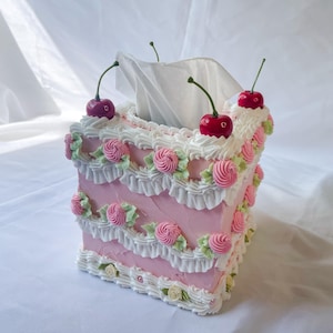 May include: A pink and white cake-shaped tissue box holder with white frosting and pink roses. The holder has three red cherries on top.