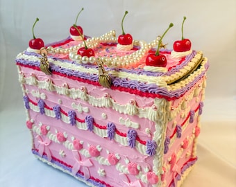 May include: A pink decorative box designed to look like a cake, decorated with white, purple, and red frosting. It is topped with red cherries, pearl strands, and gold-coloured charms. The box is decorated with pink bows and rosettes.