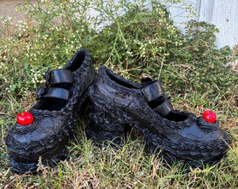 May include: A pair of black platform shoes with a textured, layered design. Each shoe features a red cherry accent and black straps. The shoes are displayed on a bed of green grass.