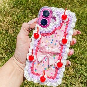 May include: A pink phone case decorated to resemble a cake. The case features white frosting, red cherries, a pink bow, and colorful sprinkles. The case is designed to fit a smartphone and has a clear camera lens opening.