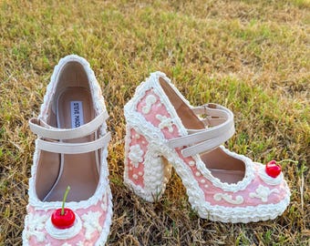 May include: Pair of whimsical pink and white high-heeled shoes with a cherry on the toe. The shoes have a cake-like design with decorative frosting and a strap across the foot. The heels are chunky and also decorated.