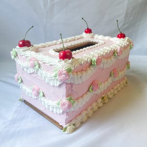 May include: A pink and white cake-shaped tissue box cover with white frosting and red cherries on top.