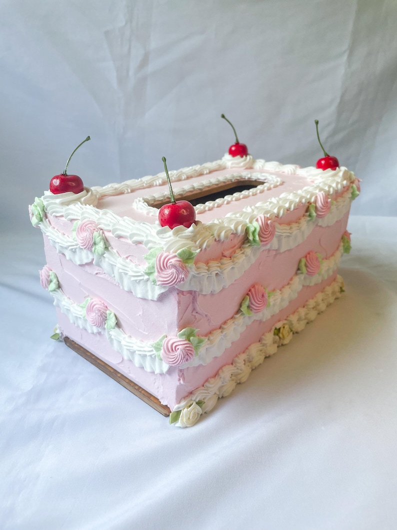 May include: A pink and white cake-shaped tissue box cover with white frosting and red cherries on top.