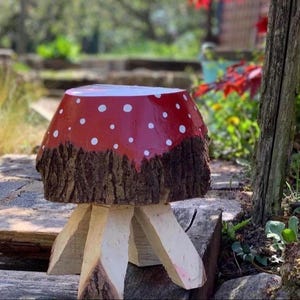 May include: A handcrafted mushroom-shaped stool. The stool has a bright red top adorned with white polka dots, a textured, dark brown base, and light-coloured wooden legs. This unique piece adds a touch of nature-inspired charm to any garden or outdoor space.