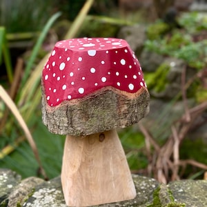 May include: A wooden mushroom ornament. The red cap features white spots, and the stem is made of natural wood. The sculpture is placed on a stone wall, with a blurred green background of plants and foliage.