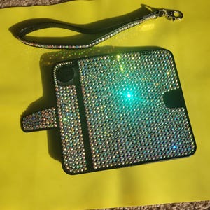May include: A black phone case with a sparkly iridescent design. The case has a flap that covers the screen and a strap.