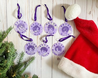 Op de afbeelding: Zeven ronde, paarse en witte ornamenten met de namen van de leden van de K-pop-groep BTS erop gedrukt. Elk ornament heeft een paars lint eraan bevestigd. De ornamenten zijn in een halve cirkel op een wit houten oppervlak gerangschikt.