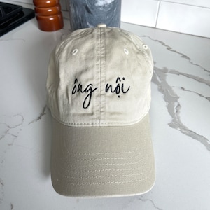May include: A beige baseball cap with a black embroidered design that says "Sông Nói".