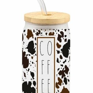 Puede incluir: Taza de cerámica blanca con un diseño de estampado de vaca marrón y negro. La taza tiene una tapa de madera y una pajita transparente. La palabra "COFFEE" está impresa en negro en la parte delantera de la taza.
