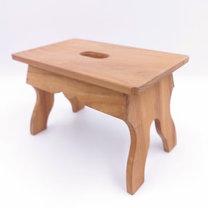 Puede incluir: Un taburete de madera con una parte superior rectangular y patas curvadas. El taburete está hecho de madera marrón claro y tiene un pequeño agujero en el centro de la parte superior.