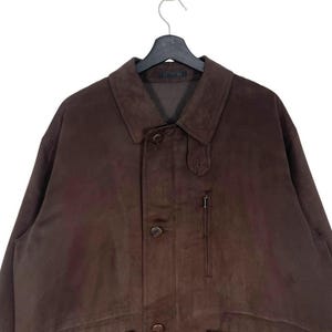 May include: A brown suede jacket with a button-up front, a collar, and a small zippered pocket. The jacket is hanging on a black hanger. The jacket appears to be a medium size.