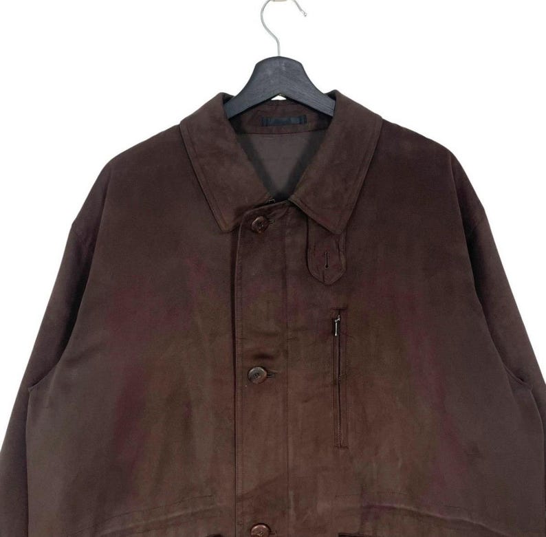 May include: A brown suede jacket with a button-up front, a collar, and a small zippered pocket. The jacket is hanging on a black hanger. The jacket appears to be a medium size.