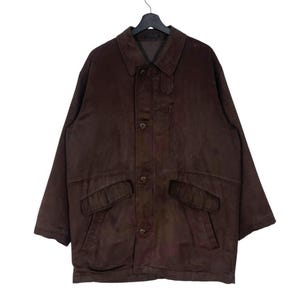 May include: A brown suede jacket with a button-up front, two front pockets, and a small chest pocket. The jacket has a collared neckline and long sleeves. It is hanging on a black hanger.