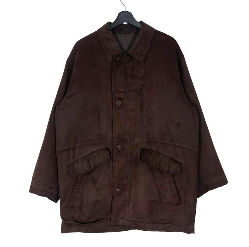 May include: A brown suede jacket with a button-up front, two front pockets, and a small chest pocket. The jacket has a collared neckline and long sleeves. It is hanging on a black hanger.