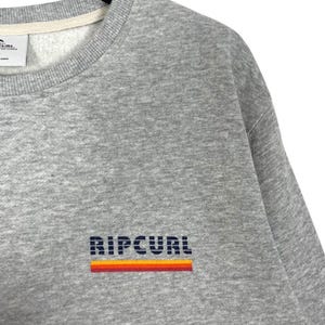 May include: A grey sweatshirt featuring the brand name "RIPCURL" in dark blue block letters with an orange and red stripe. The crew neck sweatshirt is made of a soft material. The label is visible at the top.