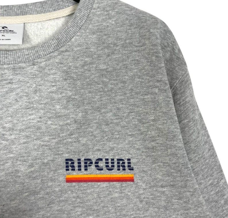 May include: A grey sweatshirt featuring the brand name "RIPCURL" in dark blue block letters with an orange and red stripe. The crew neck sweatshirt is made of a soft material. The label is visible at the top.