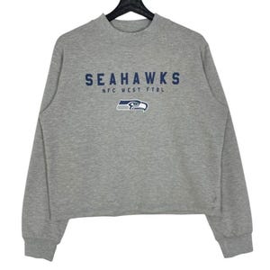 Puede incluir: Una sudadera gris con la palabra "SEAHAWKS" en letras azules oscuras en el pecho. Debajo de la palabra "SEAHAWKS" está el texto "NFC WEST FTBL." y un logotipo de un pájaro azul y blanco. La sudadera tiene cuello redondo y mangas largas.
