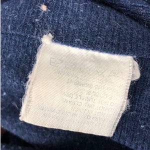 Puede incluir: Primer plano de una tela azul marino con una etiqueta rectangular blanca adjunta. La etiqueta tiene texto impreso y la tela tiene un aspecto texturizado y ligeramente borroso. La etiqueta est&aacute; sujeta a la tela con un peque&ntilde;o hilo blanco.