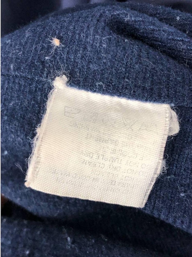 Puede incluir: Primer plano de una tela azul marino con una etiqueta rectangular blanca adjunta. La etiqueta tiene texto impreso y la tela tiene un aspecto texturizado y ligeramente borroso. La etiqueta est&aacute; sujeta a la tela con un peque&ntilde;o hilo blanco.