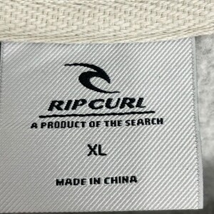 May include: A close-up of a grey fabric label with the Rip Curl logo and text. The label states "A Product of the Search", "XL", and "Made in China". The label is attached to a light beige fabric.