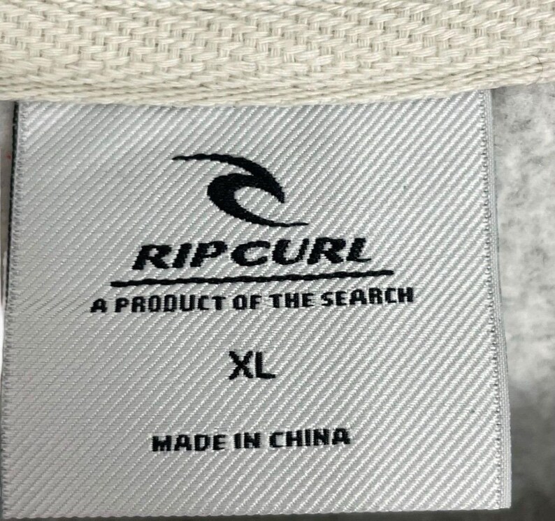 May include: A close-up of a grey fabric label with the Rip Curl logo and text. The label states "A Product of the Search", "XL", and "Made in China". The label is attached to a light beige fabric.