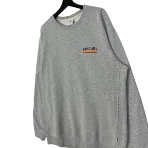 May include: A grey sweatshirt featuring the word "RIPCURL" in orange, red, and blue across the chest. The sweatshirt has a crew neck and long sleeves. The garment is displayed on a black hanger.