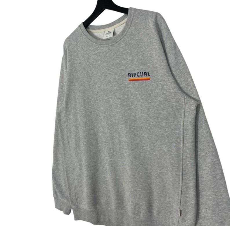 May include: A grey sweatshirt featuring the word "RIPCURL" in orange, red, and blue across the chest. The sweatshirt has a crew neck and long sleeves. The garment is displayed on a black hanger.