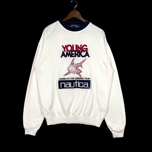 以下が含まれることがあります： ネイビーブルーの襟と袖口が付いた白い Nautica スウェットシャツ。「YOUNG AMERICA」の赤い文字、赤と白のグラフィック、ネイビーブルーの長方形に「nautica」の文字が特徴です。 グラフィックの下には「AMERICA'S CUP DEFENSE TEAM」と印刷されています。
