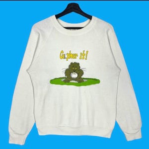 May include: White crew neck sweatshirt with a cartoon gopher graphic holding a golf ball. The text "Gopher it!" is above the gopher. The gopher is green and brown, and the text is yellow.