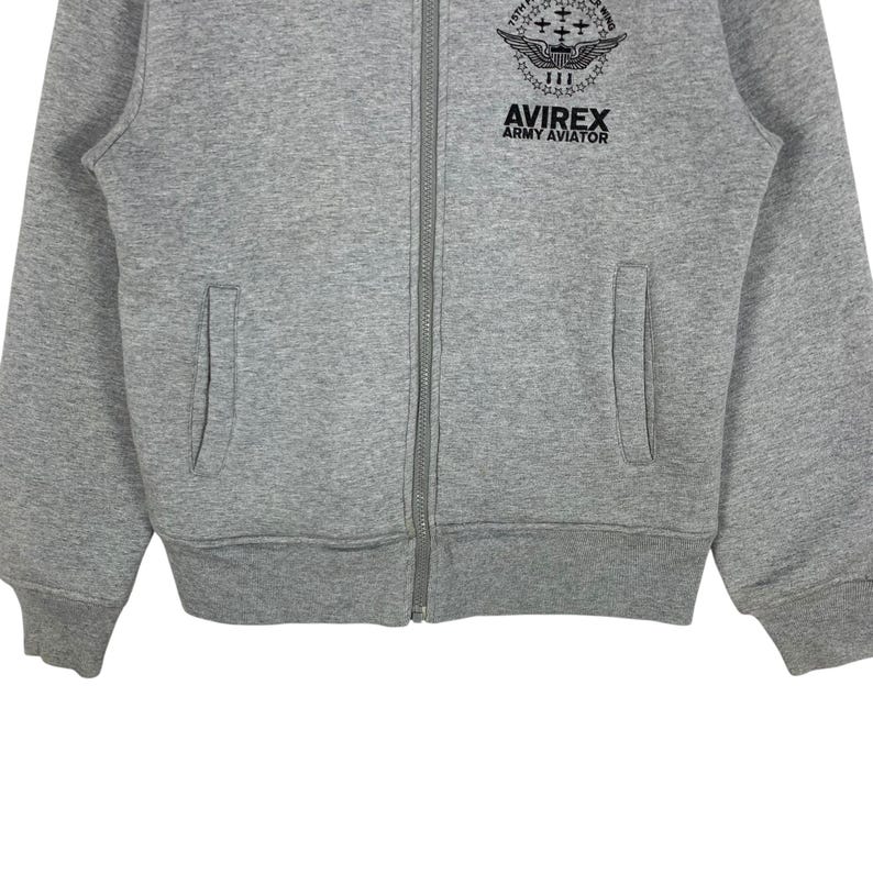 May include: A gray zip-up sweatshirt with two front pockets. The sweatshirt features the text "AVIREX ARMY AVIATOR" and a graphic on the upper chest. The jacket has a full-length zipper and ribbed cuffs and hem.