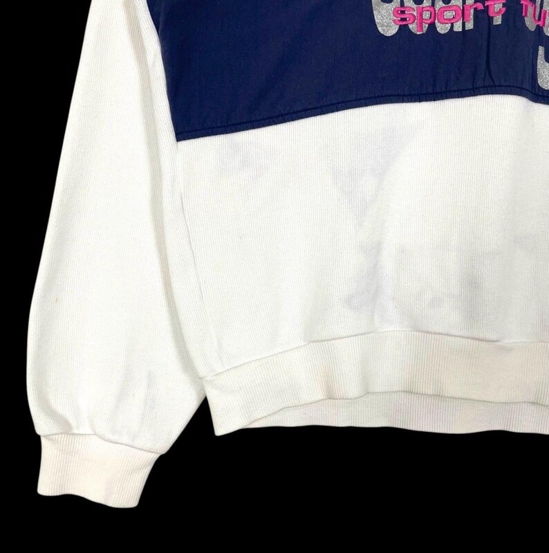 Puede incluir: Sudadera blanca con un panel superior azul marino. El panel superior tiene el texto "sport" en letras rosas y plateadas. La sudadera tiene pu&ntilde;os y dobladillo acanalados.