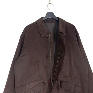 May include: A brown suede jacket with a collared neckline, button closures, and two front pockets. The jacket is hanging on a black hanger against a white background. The jacket appears to be a vintage or pre-owned item.